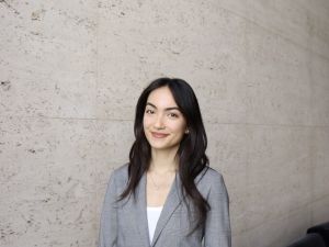 A picture of Sera Balkir in a grey blazer, standing in front of a light grey concrete wall.